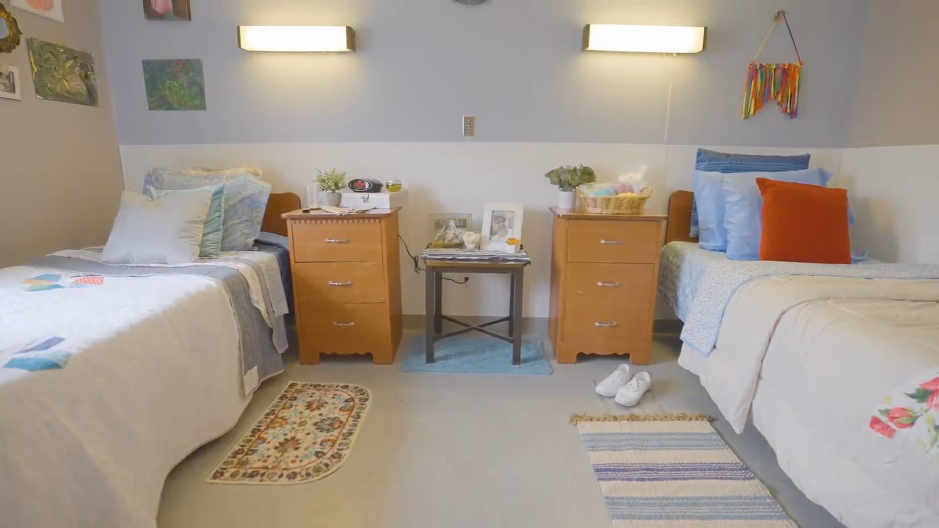 A shared bedroom with two twin beds separated by wooden nightstands, wall-mounted lights, rugs, and personal decor.