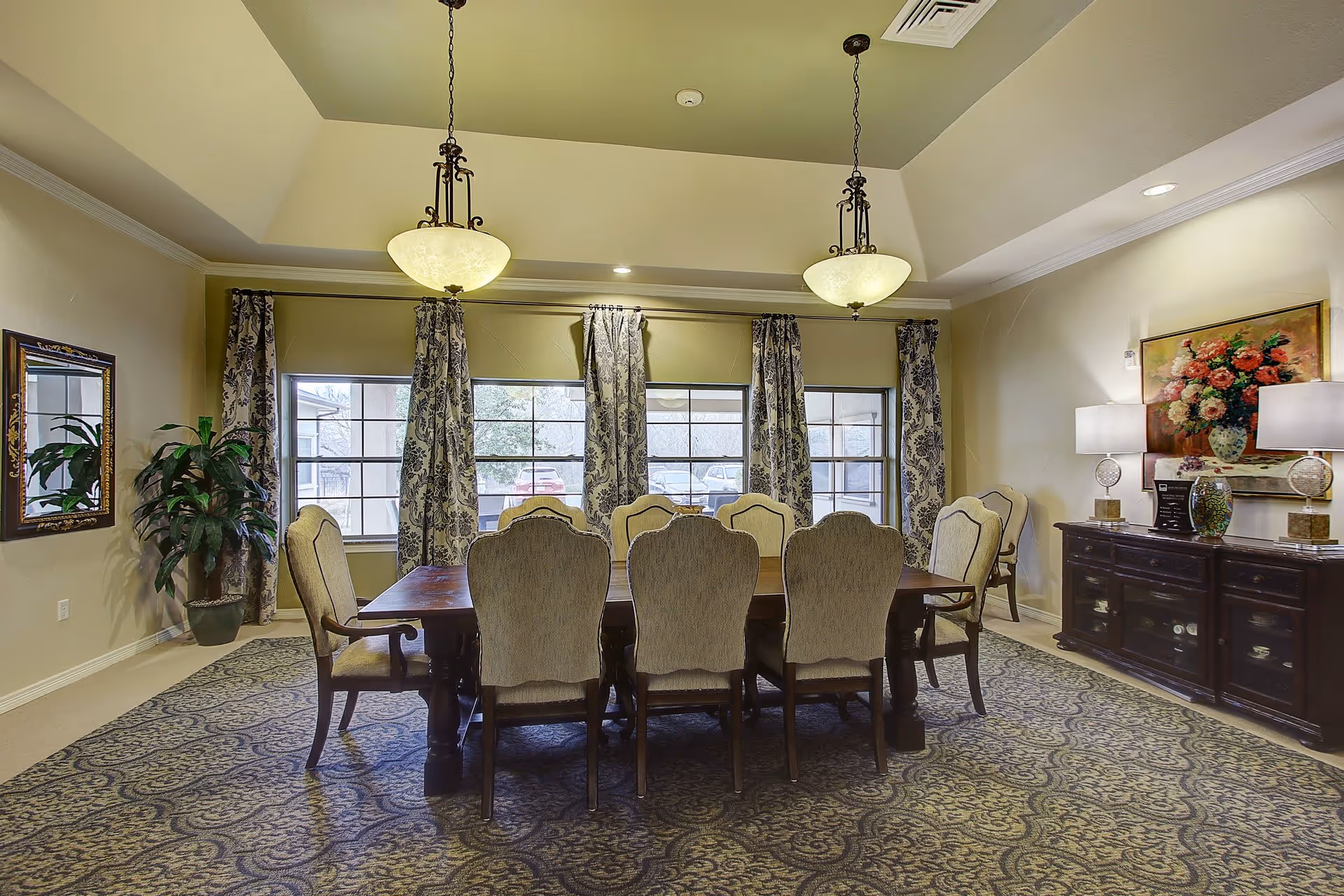 A formal dining room with a large wooden dining table surrounded by eight upholstered chairs. The room features patterned curtains on three large windows, two hanging pendant lights, a decorative rug, a potted plant, a mirror on one wall, and a dark wooden sideboard with two table lamps and a floral painting above it.