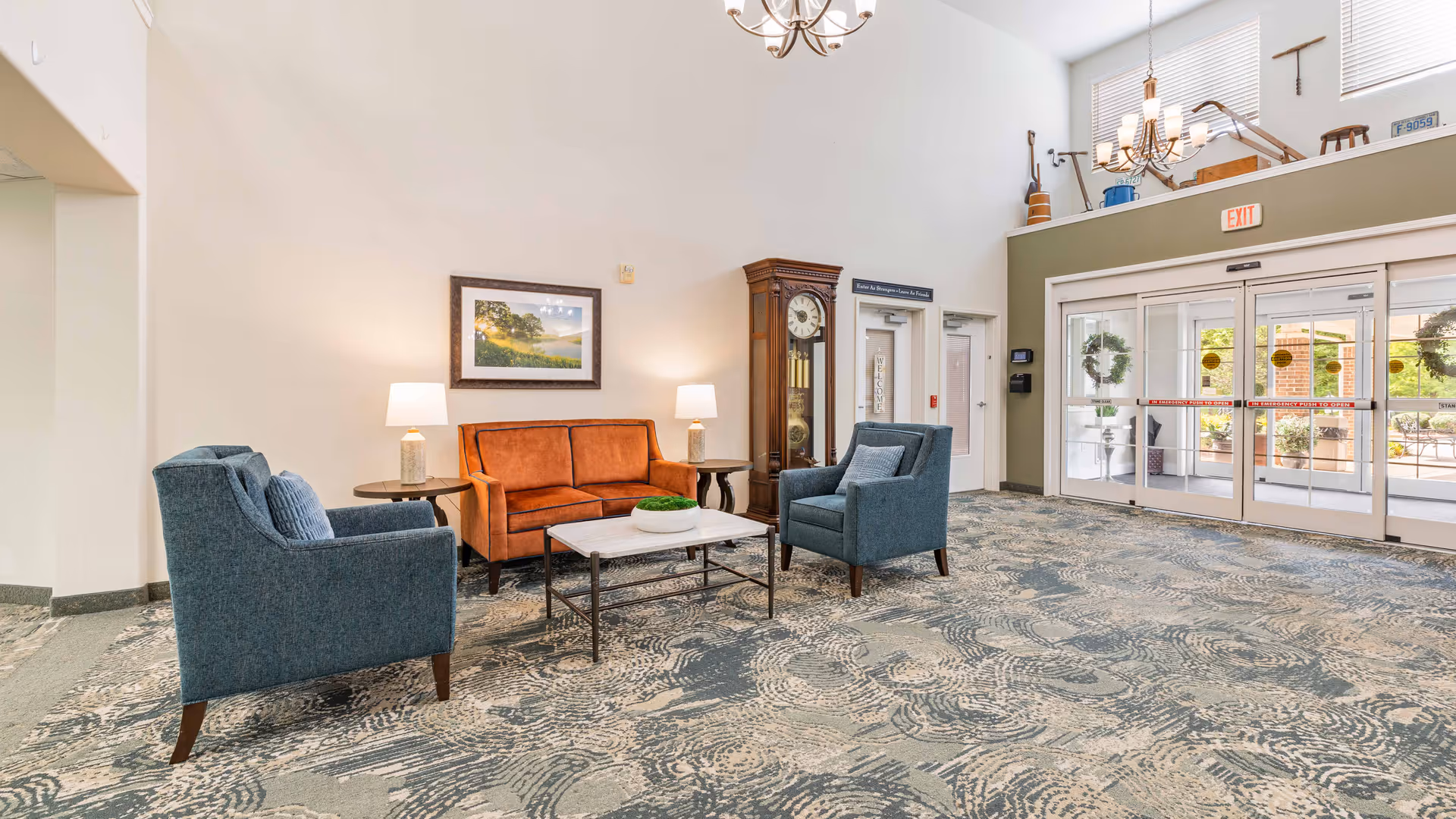 A spacious and well-lit retirement facility lobby with patterned carpet flooring, a seating area consisting of an orange sofa and two blue armchairs around a coffee table, two table lamps on side tables, a large grandfather clock, and glass double doors leading outside. The walls are light-colored, and there is a framed landscape picture above the sofa. The ceiling is high with chandeliers and a loft area above the entrance.