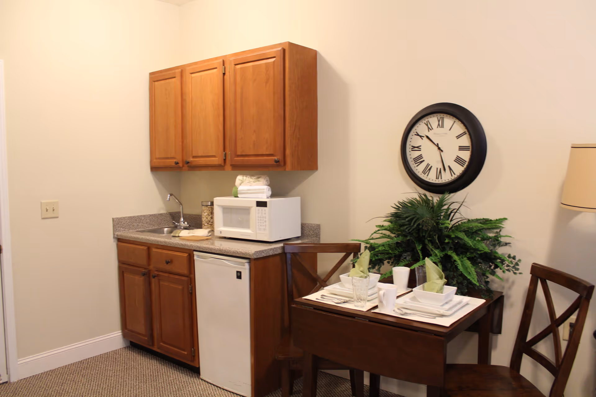 Small kitchenette area with wooden cabinets, a countertop with a sink, a microwave, and a mini refrigerator. Next to the kitchenette is a small wooden dining table set for two with white dishes, green napkins, and a plant centerpiece. A round wall clock with Roman numerals hangs above the table, and a floor lamp is partially visible on the right.
