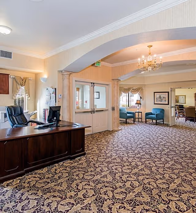Interior view of a senior living facility reception area with a dark wooden desk and black office chair on the left. The room has patterned carpet, beige walls, and an arched doorway leading to a sitting area with blue chairs and a small table under a chandelier. Double doors with glass panels are visible in the center background.
