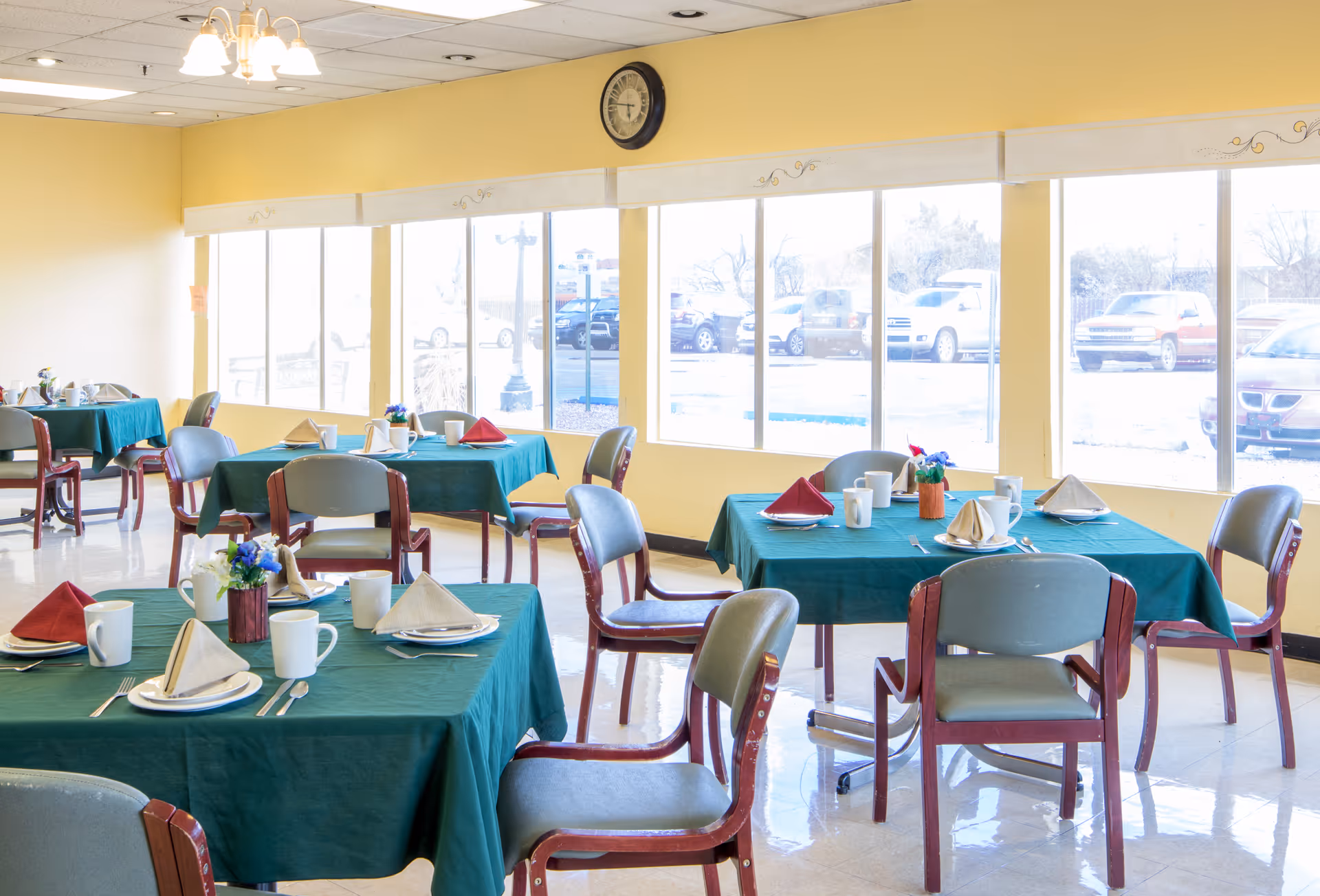 A bright dining room with several tables covered in green tablecloths, each set with plates, cups, folded napkins, and silverware. Chairs with wooden frames and gray cushions surround the tables. Large windows along one wall let in natural light and show parked cars outside. A clock hangs above the windows on a yellow wall.