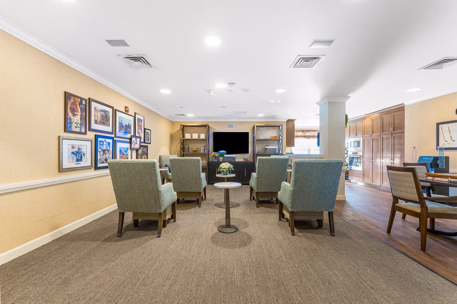 Bright communal lounge with several upholstered chairs facing a TV and shelving, framed photos on the left wall, and a dining/kitchen area visible to the right.