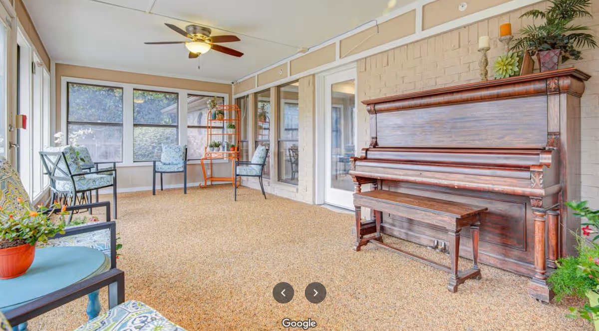 A bright enclosed patio area with large windows, featuring several cushioned chairs with floral patterns, a small round table with a potted plant, an antique wooden upright piano with a matching bench, and decorative items on top of the piano. The ceiling has a fan with a light fixture.