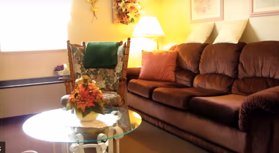 Cozy living room with a brown upholstered sofa, a floral armchair, a glass coffee table topped with a flower arrangement, and a lit floor lamp.