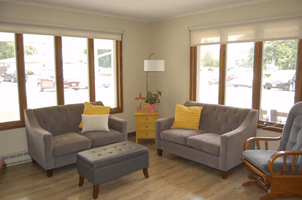 A cozy living room with two gray tufted sofas, each with yellow and white pillows, a gray tufted ottoman, a wooden rocking chair with gray cushions, a small yellow side table with a plant and a cross, and large windows letting in natural light.