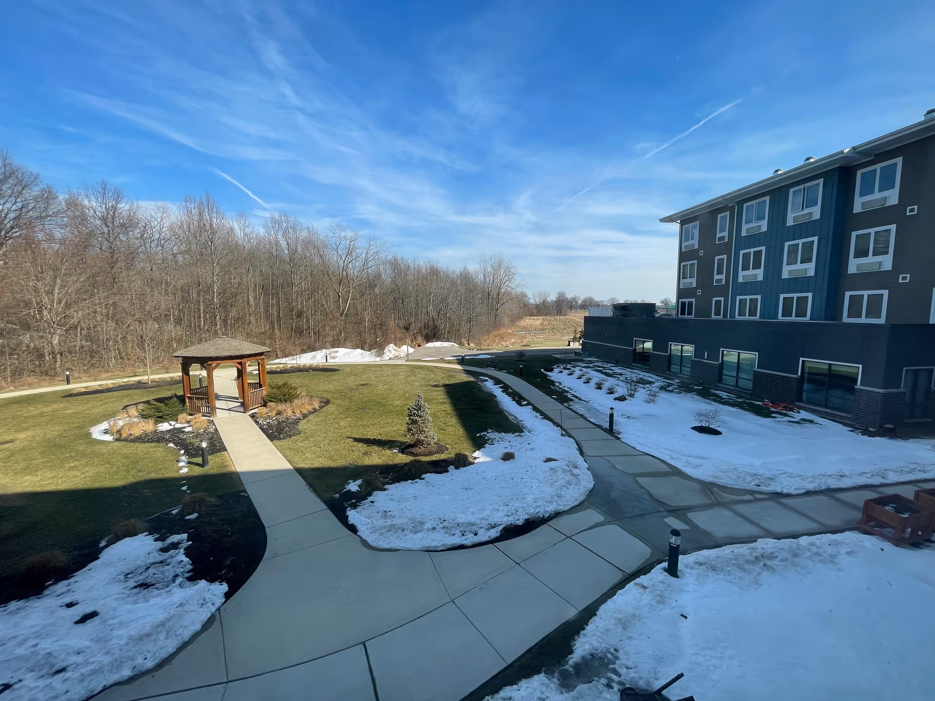 Outdoor view of a senior living facility with a paved walkway leading to a wooden gazebo surrounded by grass and patches of snow. The building is visible on the right side, and leafless trees are in the background under a clear blue sky.