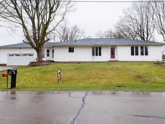 Single-story white house with a gray roof, a large front lawn, a tree on the left side, a mailbox near the street, and a 'For Sale' sign on the grass. The house has a garage on the left and a purple front door.