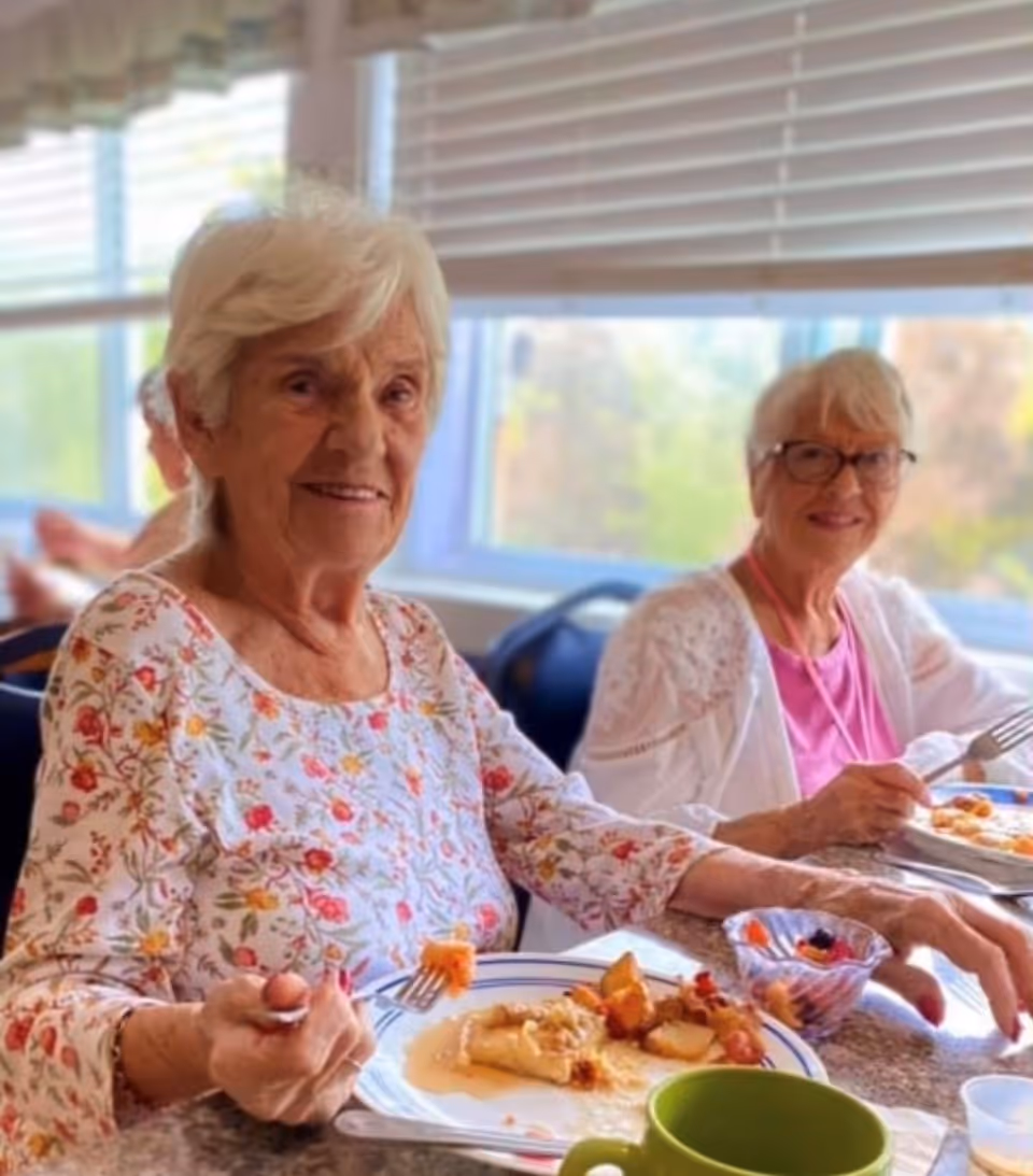 Two elderly women sitting at a dining table enjoying a meal together in a bright room with large windows and blinds.