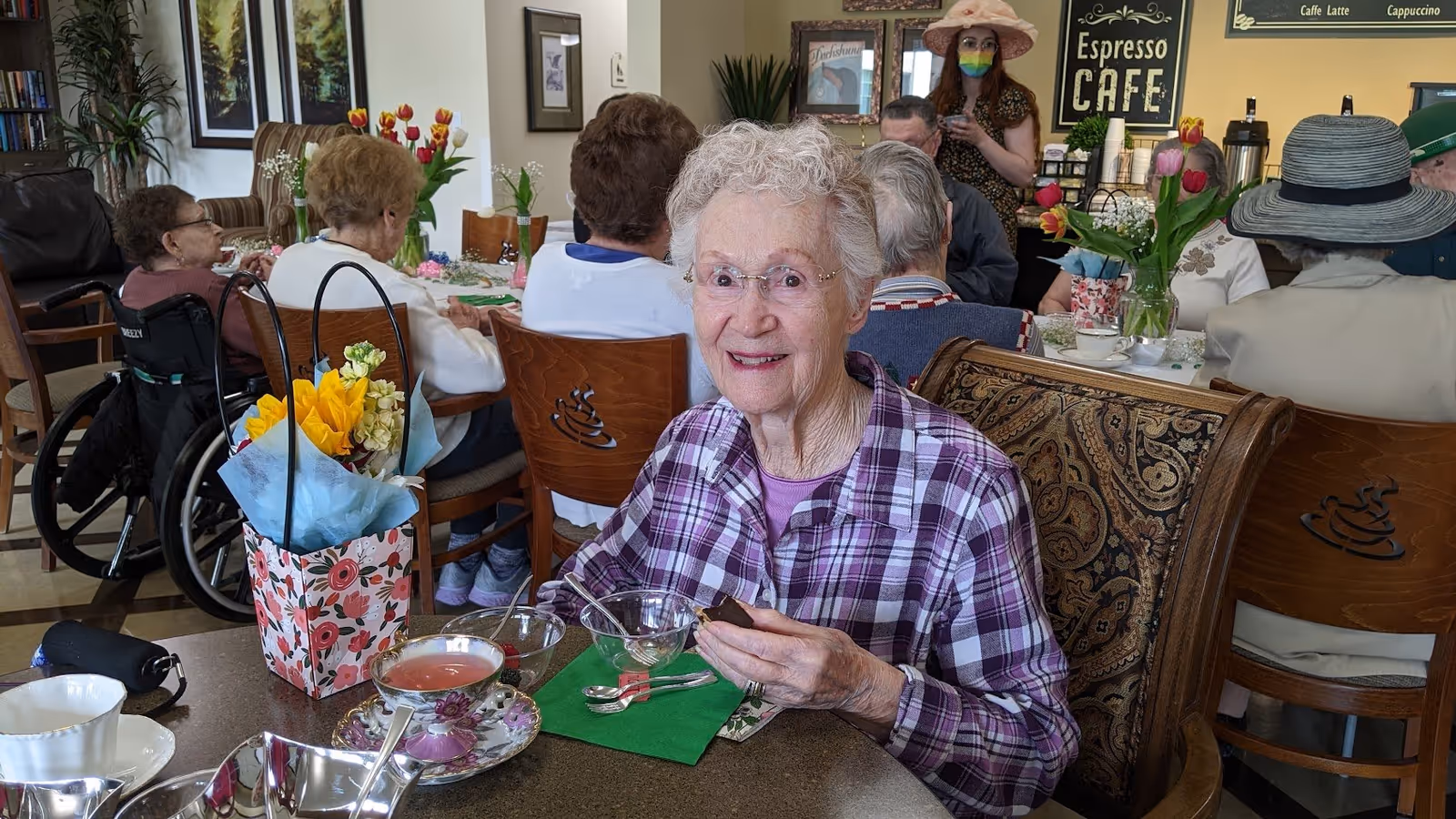 An older woman in a plaid shirt smiling while seated at a table with tea, flowers, and dishes in a communal dining room.