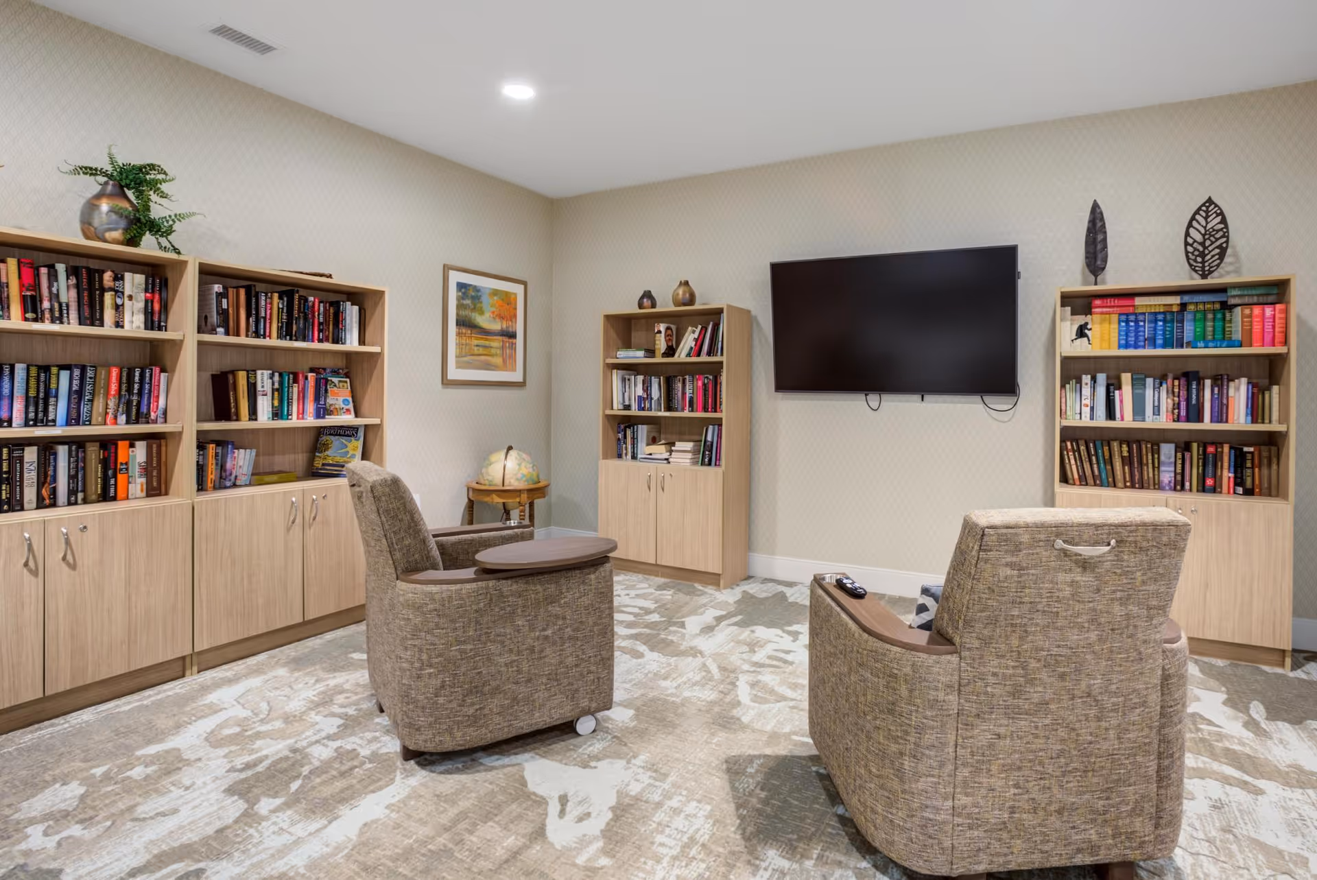 A cozy reading and relaxation room with three wooden bookshelves filled with books, two comfortable upholstered chairs with attached side tables facing a wall-mounted flat screen TV, a globe on a small stand, and a framed landscape painting on the wall. The room has a patterned carpet and soft lighting.