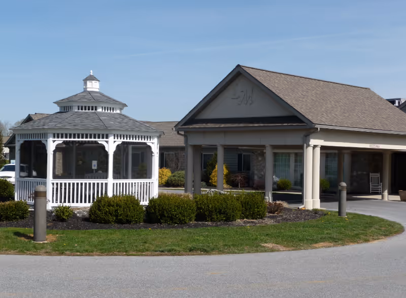 Exterior view of a senior living facility featuring a white gazebo surrounded by bushes and a covered entrance with columns and a peaked roof under a clear blue sky.