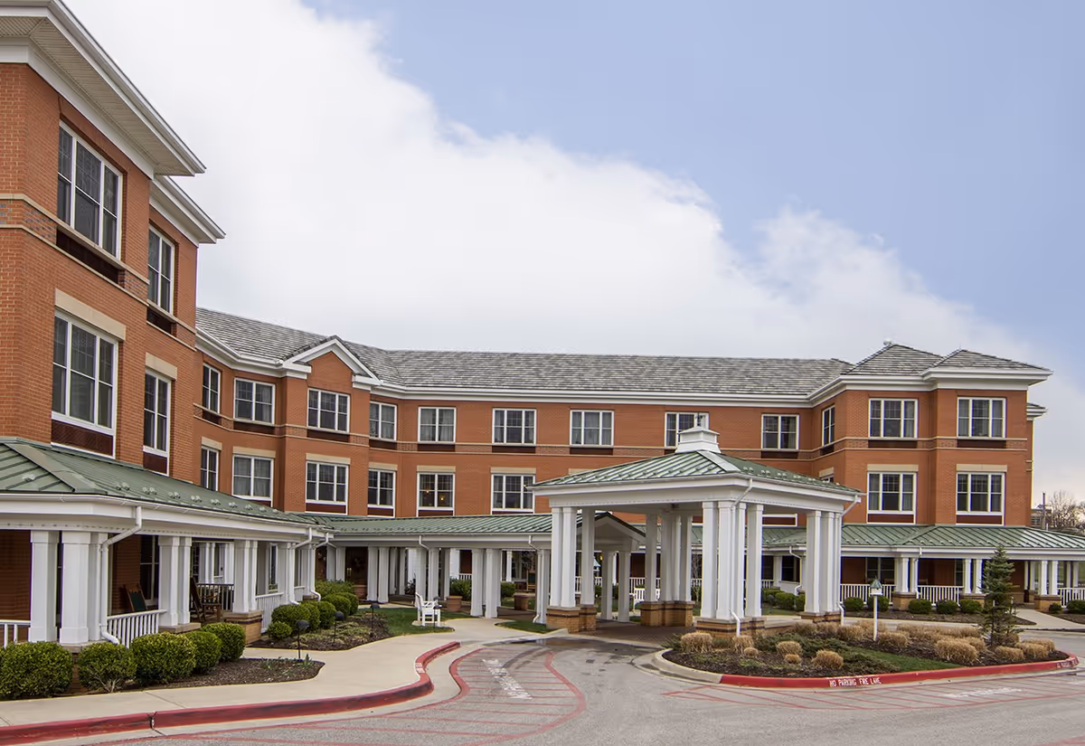 Exterior view of a multi-story brick building with many windows, a covered entrance with white columns, and a driveway with a red curb. The sky is partly cloudy.