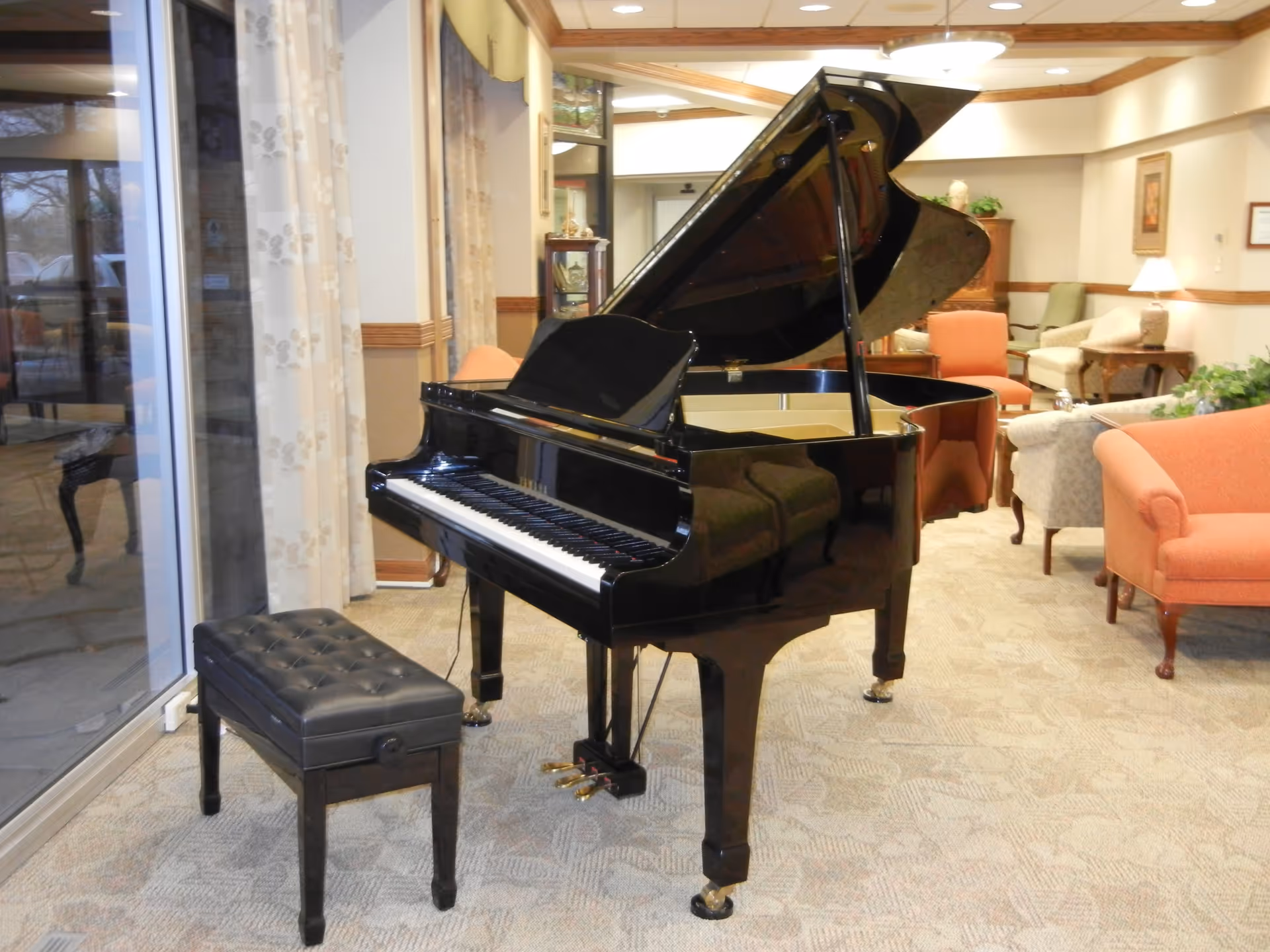 A black grand piano with an open lid and a black cushioned bench in a carpeted room. The room has large windows with floral curtains on the left, and various upholstered chairs and sofas in soft colors arranged around the space. The walls are light-colored with wooden trim, and there are framed pictures and lamps in the background.
