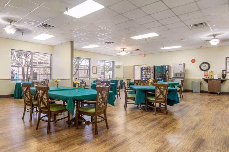 Spacious dining/activity room with round tables covered in green tablecloths, chairs, windows, and a vending/beverage station.