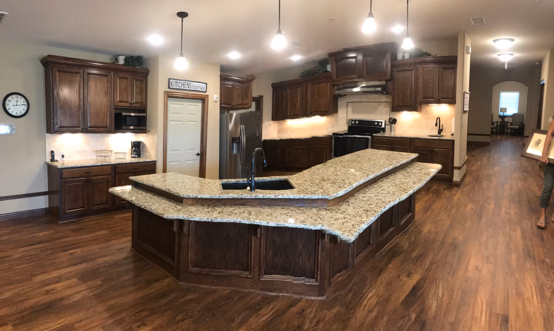 A spacious kitchen with dark wooden cabinets and a large granite island countertop with a built-in sink. The kitchen features stainless steel appliances including a refrigerator and stove. The floor is wooden, and there are pendant lights hanging from the ceiling. A hallway is visible in the background, and a person is partially visible on the right side holding a framed picture.
