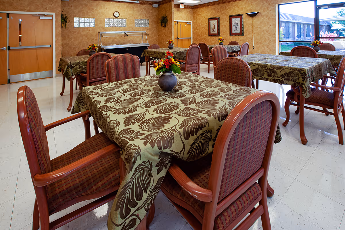 A dining room with several tables covered in green and brown patterned tablecloths. Each table has a small vase with colorful flowers. The room has wooden chairs with red cushioned seats and backs. There are framed pictures on the walls, a clock above a door, and large windows letting in natural light.