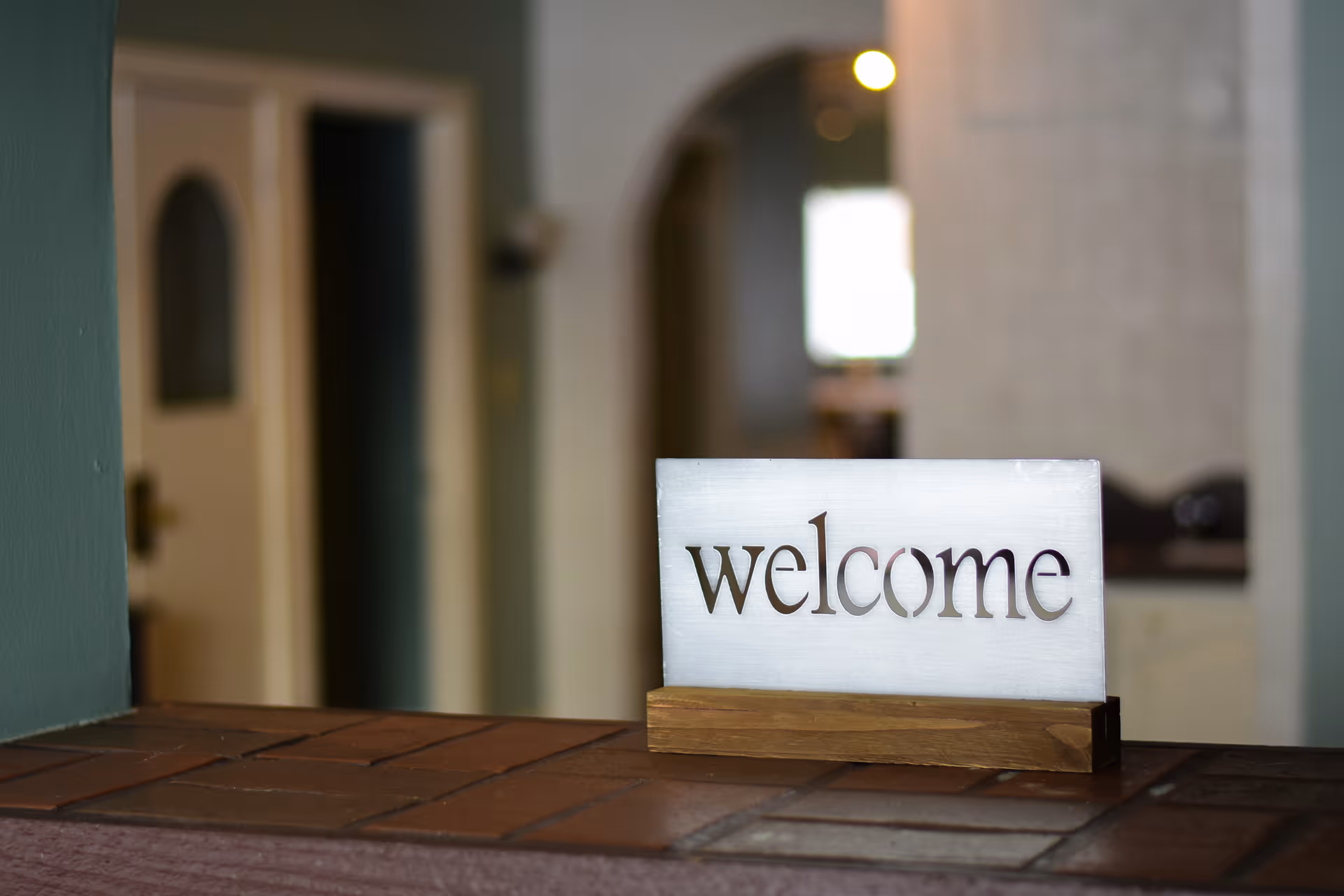 A close-up of a wooden and metal sign with the word 'welcome' cut out, placed on a tiled counter with a blurred background of an interior hallway and door.