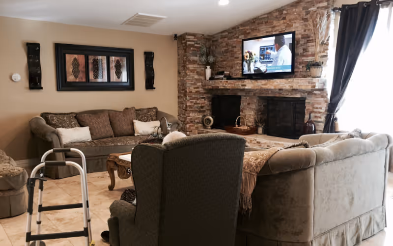 A cozy living room in an assisted living facility with a stone fireplace, a wall-mounted TV, multiple cushioned sofas and armchairs, a coffee table, decorative wall art, and a walker placed near the seating area. Natural light comes through a window with dark curtains.