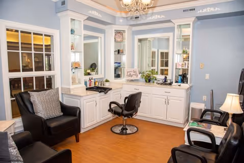 A well-lit salon area with white cabinetry and large mirrors, featuring a black salon chair in front of the counter. There are three black chairs with cushions arranged around the room, a small lamp on a side table, and various hair care products on the counter. The walls are painted light blue with white trim, and the floor is wooden.