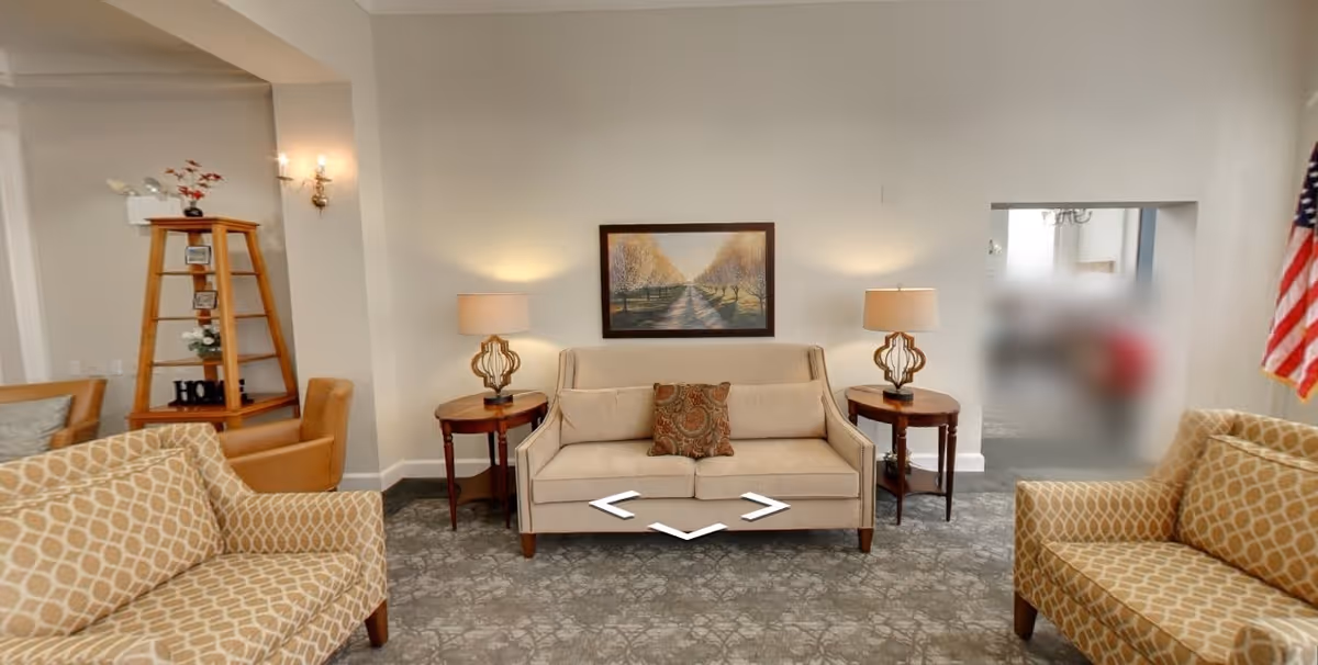 A cozy living room area with a beige sofa centered against a white wall, flanked by two wooden side tables each with a decorative lamp. Above the sofa hangs a framed painting of a tree-lined path. Two patterned armchairs face the sofa, and a wooden ladder-style shelf with decorative items is visible to the left. An American flag is partially visible on the right side of the image.