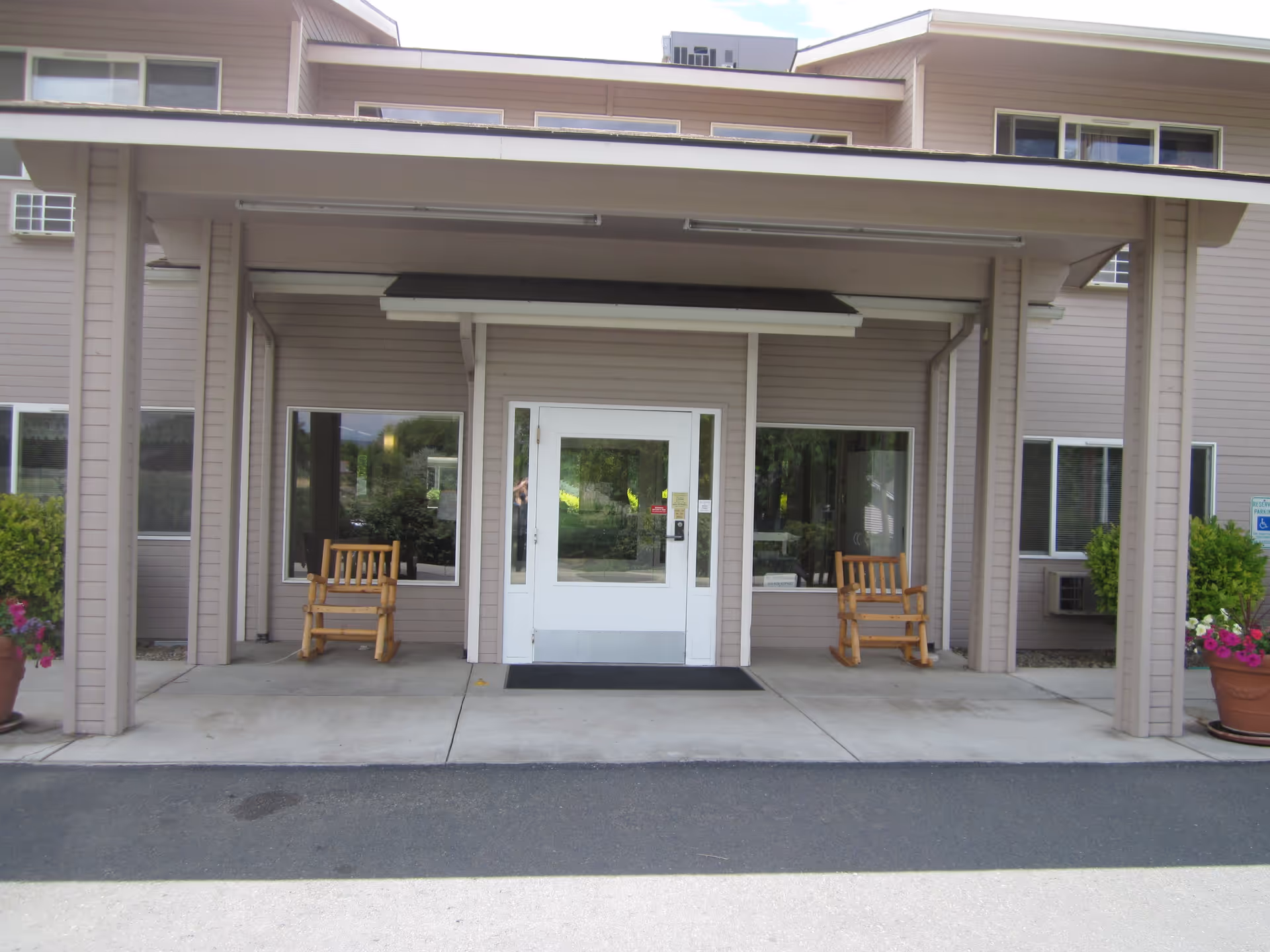 Entrance to Sunnyside Assisted Living facility with a covered porch, two wooden rocking chairs on either side of a white door, large windows, potted plants with flowers, and beige siding on the building.