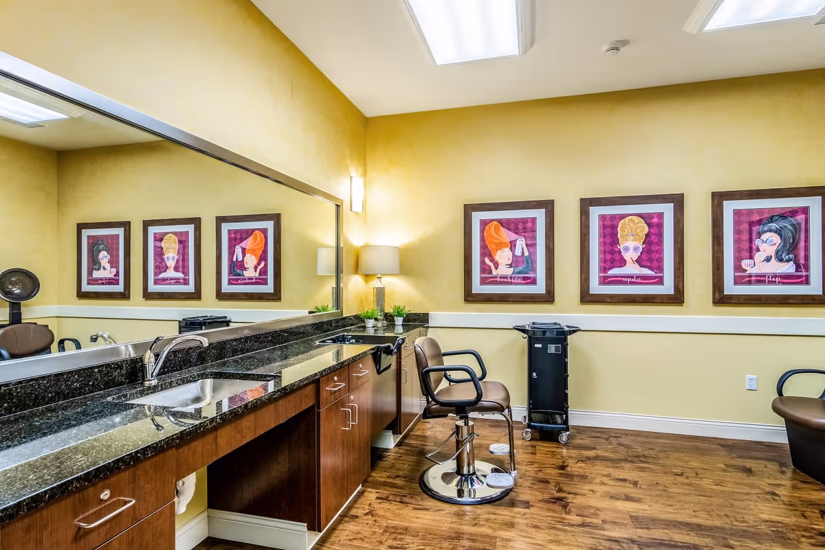 Interior of a salon room with a large mirror above a granite countertop, a sink, wooden cabinets, a salon chair, a rolling cart, and three framed colorful caricature-style portraits on a yellow wall.