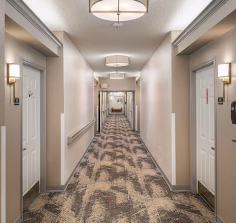 A long, well-lit hallway in a senior living facility with patterned carpet, beige walls, white doors on both sides, wall-mounted lights, and ceiling light fixtures. There is a handrail along the left wall and a bench visible at the far end of the hallway.
