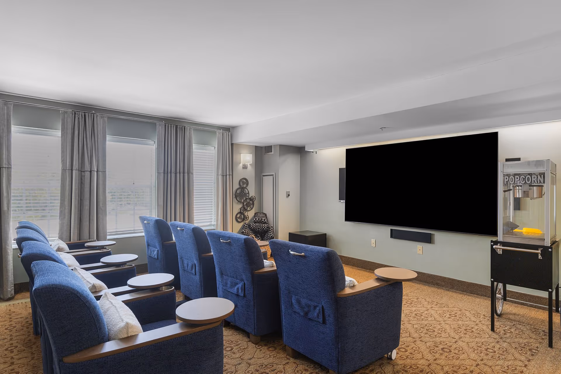 A small theater-style room with two rows of blue upholstered recliner chairs, each with a small round table attached. The room has large windows with gray curtains on the left side, a large flat-screen TV mounted on the wall, and a popcorn machine on the right side. The carpet has a patterned design, and the walls are painted light gray.