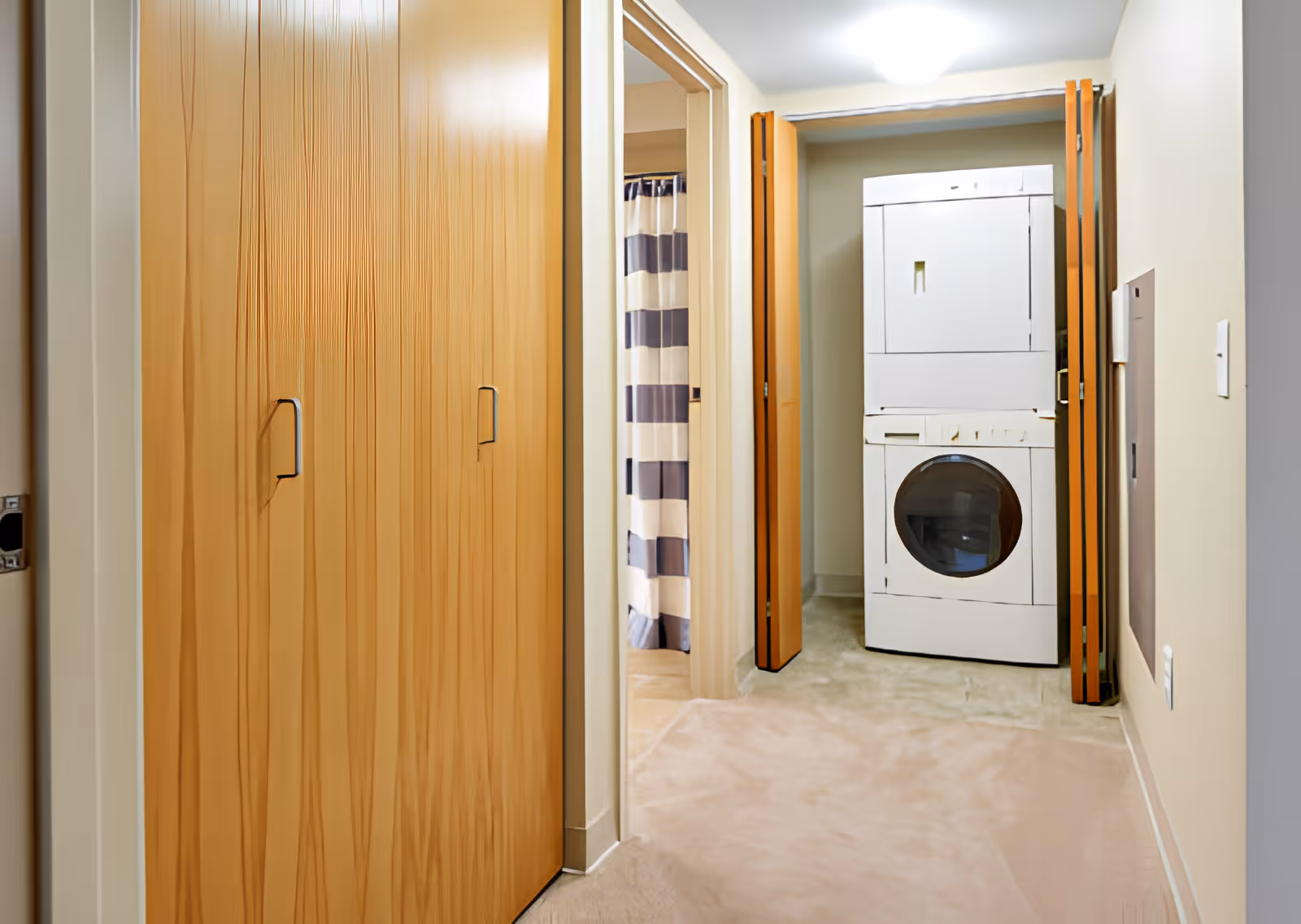A hallway with wooden closet doors on the left and a laundry area with a stacked washer and dryer at the end. To the left of the laundry area, there is a bathroom with a shower curtain featuring horizontal stripes in white and dark blue.