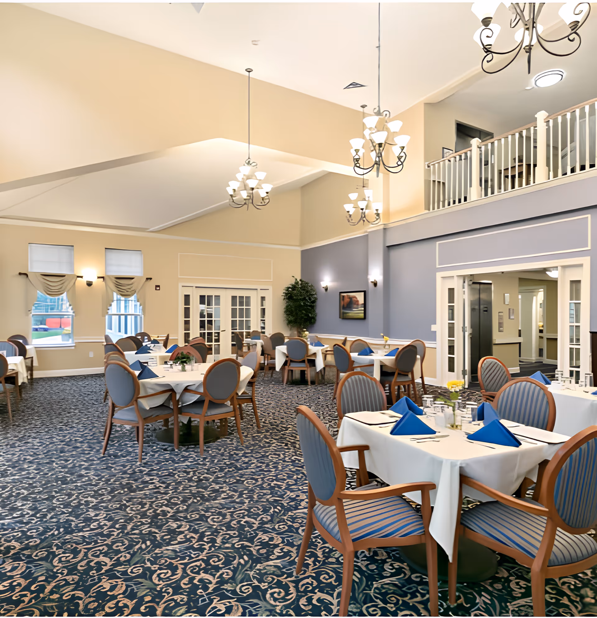 A spacious dining room in an assisted living facility with multiple round tables covered with white tablecloths and set with blue folded napkins, glasses, and silverware. The room features patterned carpet, beige and blue walls, large windows with drapes, and several chandeliers hanging from a high ceiling. There is a balcony railing visible on the upper right side and a large plant near the back wall.