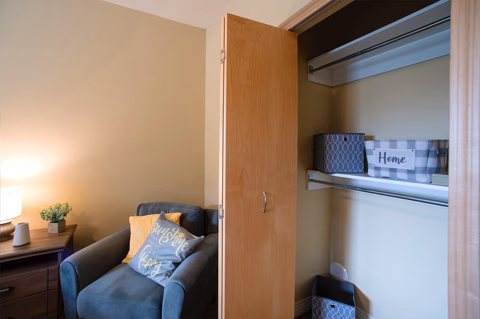 A cozy corner of a room featuring a blue armchair with two decorative pillows, one yellow and one gray with text. Next to the chair is a wooden nightstand with a lamp and a small plant. An open wooden closet door reveals shelves with storage bins, one labeled 'Home'.