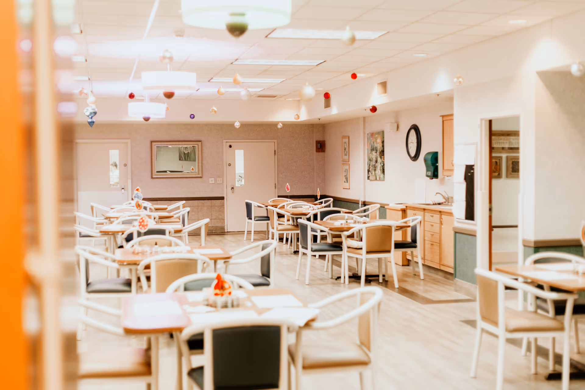 A bright dining room in a senior living community with several tables and chairs arranged neatly. The room features light-colored walls, hanging ceiling lights, and small decorative ornaments suspended from the ceiling. There are doors and framed artwork on the walls, and a clock and a hand sanitizer dispenser are visible near a counter area.