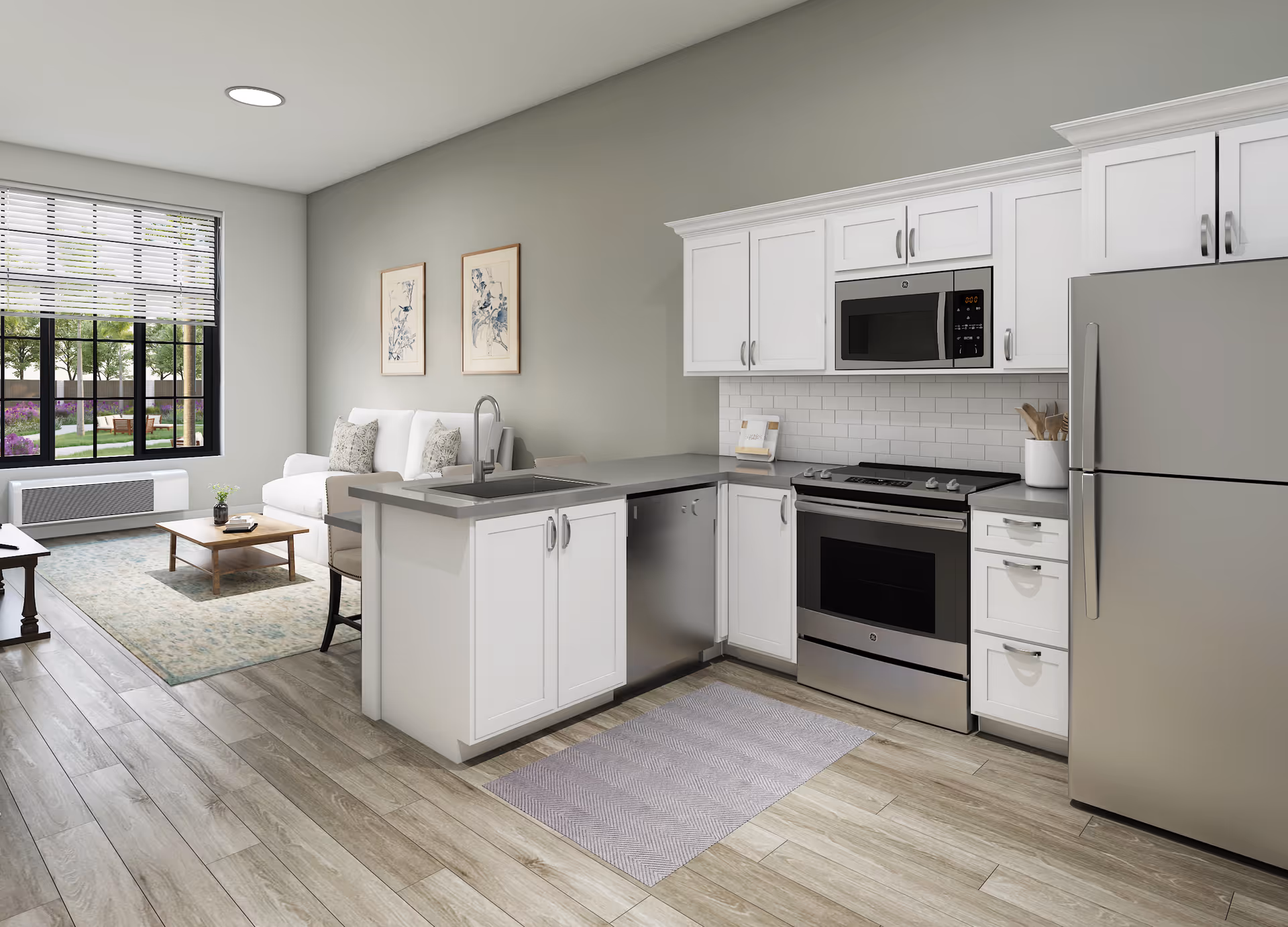 Modern open-concept kitchen and living room area with white cabinetry, stainless steel appliances including a refrigerator, oven, microwave, and dishwasher. The kitchen has a gray countertop and a small rug on the wooden floor. The living room features a white sofa, a wooden coffee table, two framed artworks on the wall, and a large window with blinds overlooking a garden outside.