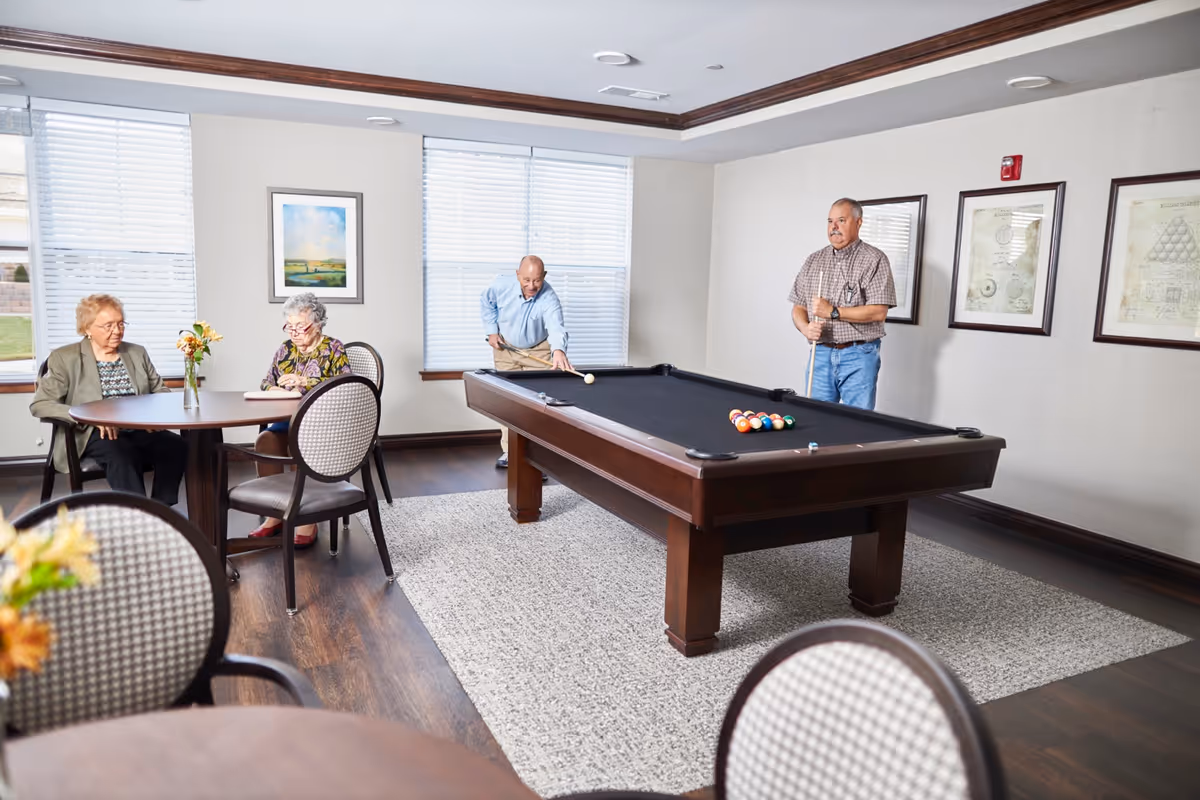 A bright room with two elderly men playing pool on a black felt pool table while two elderly women sit at a round table nearby, one reading a book. The room has large windows with blinds, framed artwork on the walls, and wooden flooring with a light-colored rug under the pool table.