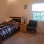A simple bedroom with a single bed covered in a blue and white checkered blanket, a pillow, a wooden nightstand with a lamp, a black cushioned chair, and a window with blinds partially open letting in natural light.