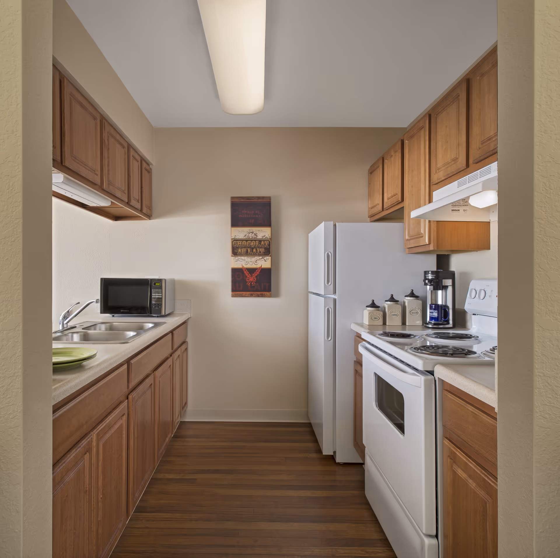 A small kitchen with wooden cabinets, a white refrigerator, white electric stove with oven, a microwave on the counter, a coffee maker, and three decorative canisters. There is a double sink on the left side and a wall decoration hanging on the back wall. The floor is wooden and the walls are painted beige.