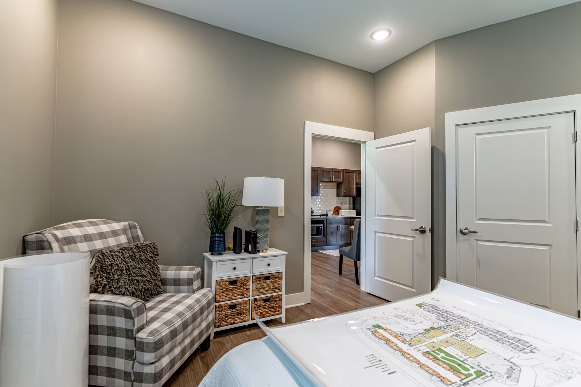 A cozy interior room with a checkered armchair, a small white cabinet with wicker baskets, a table lamp, and a potted plant. The room has beige walls and wood flooring. Two white doors are visible, one open showing a kitchen area with dark cabinets and a dining chair.