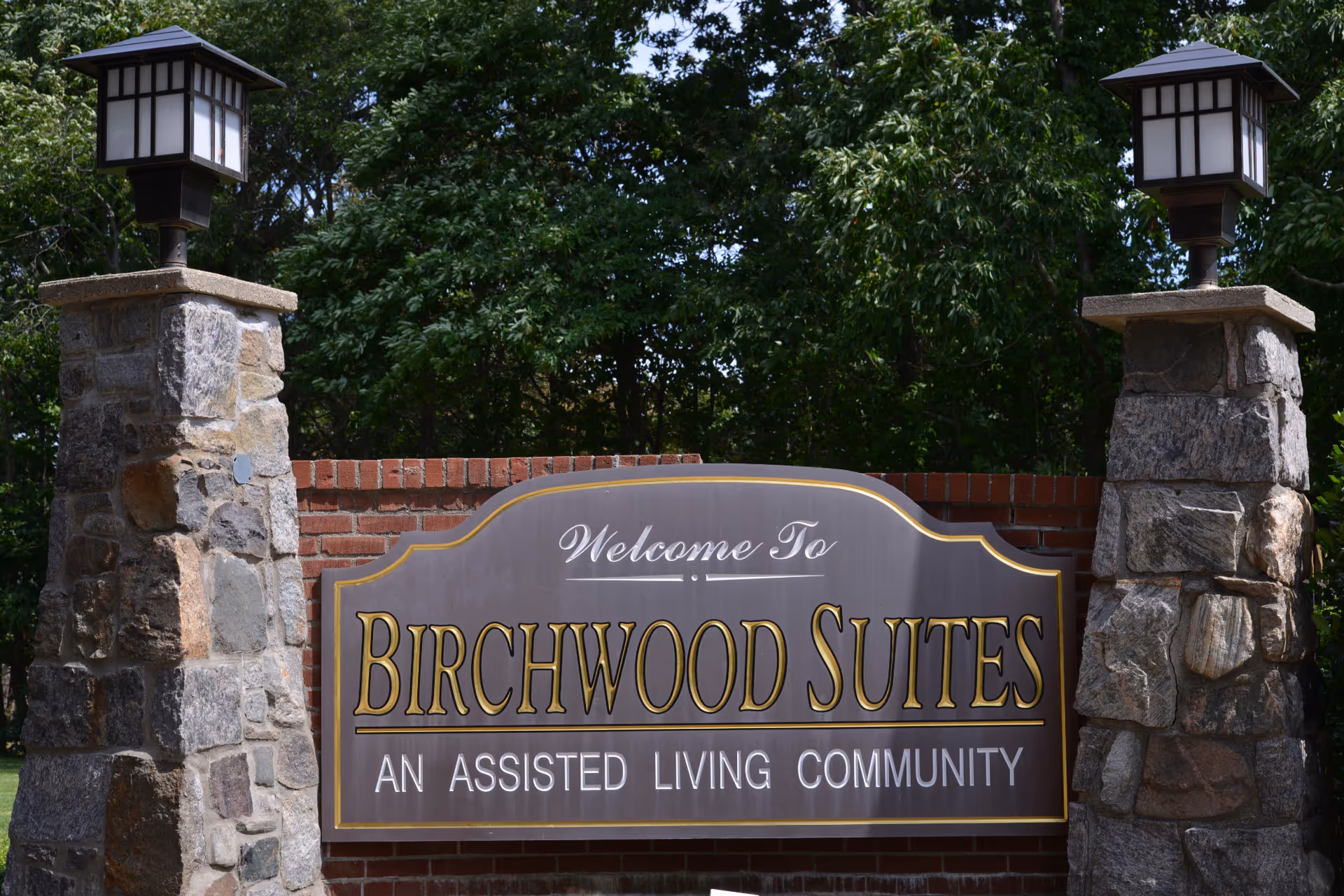 A stone and brick entrance sign for Birchwood Suites, an assisted living community, flanked by two stone pillars topped with lantern-style lights, with green trees in the background.