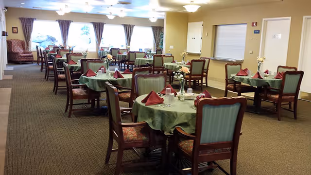 A dining room with multiple round tables covered with green tablecloths and set with red folded napkins, glasses, and small flower arrangements. The room has carpeted floors, large windows with curtains, and several wooden chairs with cushioned seats and backs. The lighting is provided by ceiling fixtures.