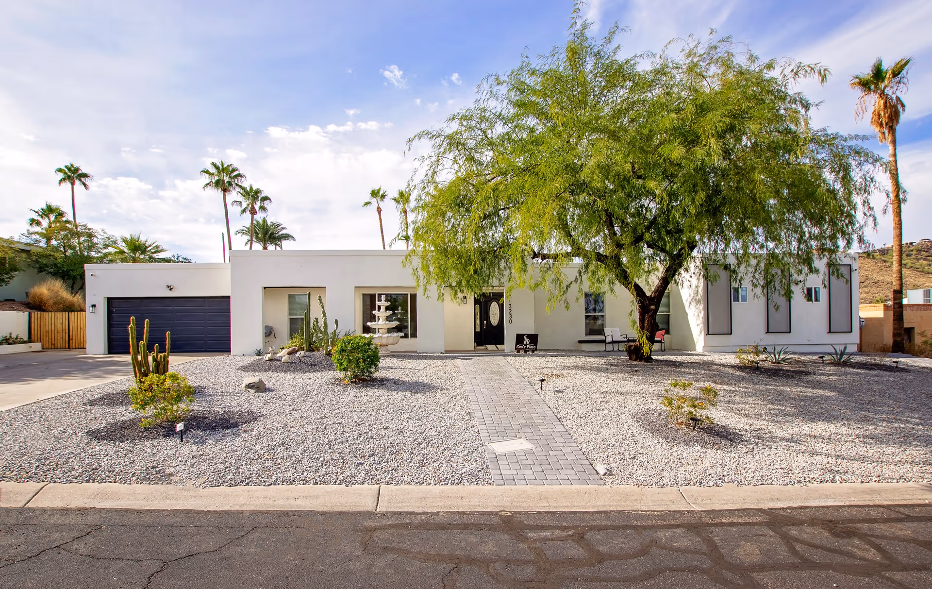 Front exterior view of a single-story white building with a flat roof, a large green tree in the front yard, desert landscaping with gravel and small plants, a paved walkway leading to the entrance, and a two-car garage with a dark door on the left side. The sky is partly cloudy with palm trees in the background.