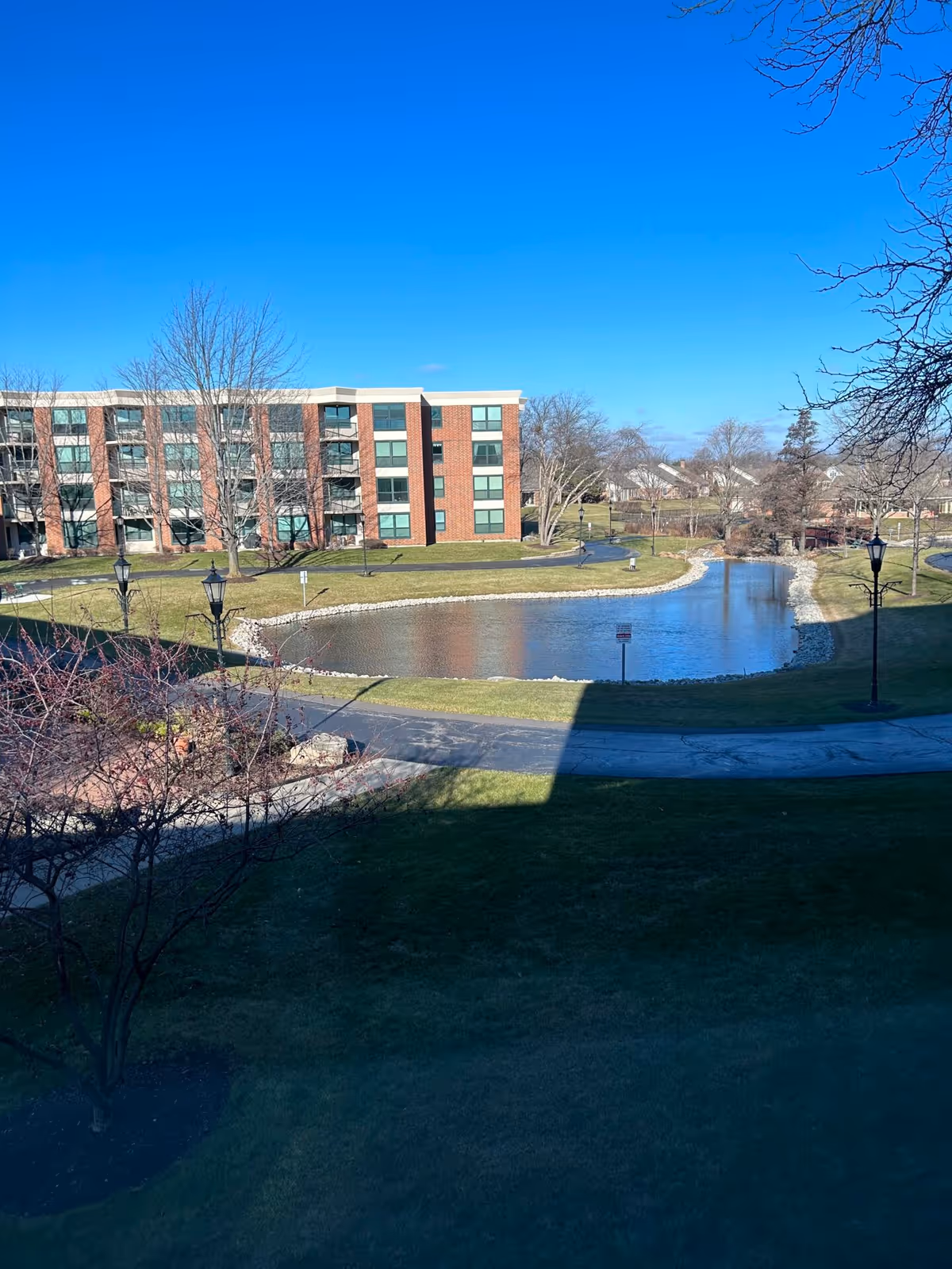 A brick multi-story residential building overlooking a landscaped pond and pathways under a clear blue sky.