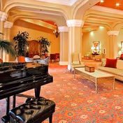 Elegant interior room with a black grand piano and cushioned bench in the foreground, ornate columns, patterned carpet, and comfortable seating including sofas and chairs arranged around a coffee table. The room is warmly lit with chandeliers and decorated with plants and framed artwork.