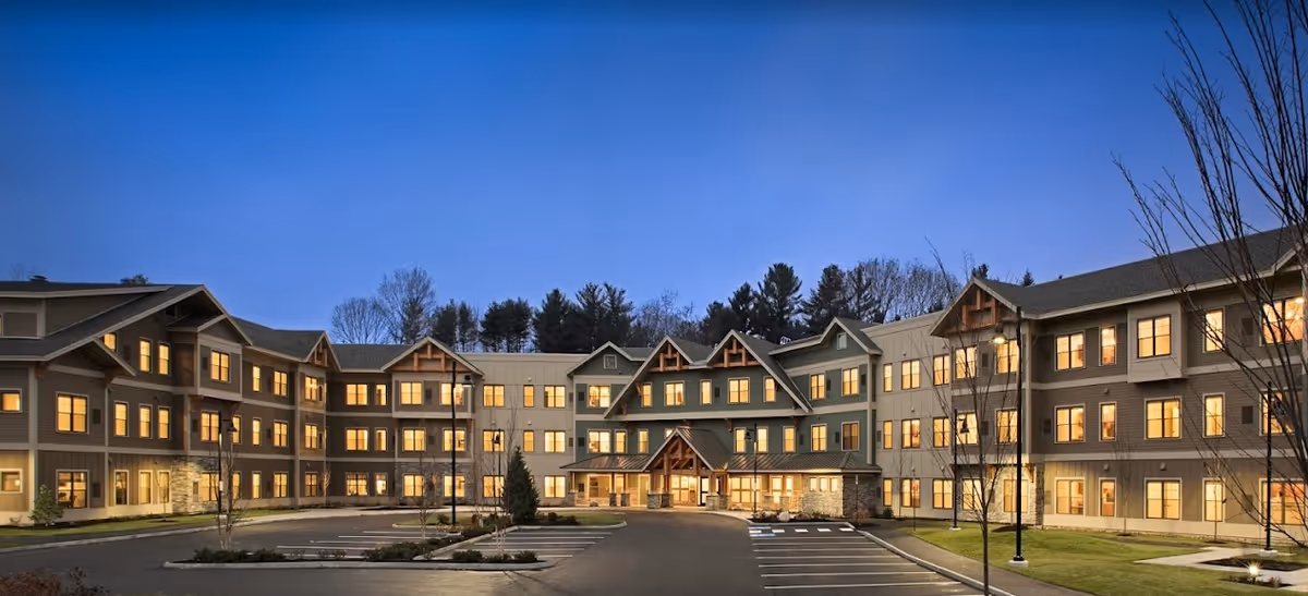 Front exterior of a multi-story senior living building at dusk with illuminated windows and an empty parking lot.