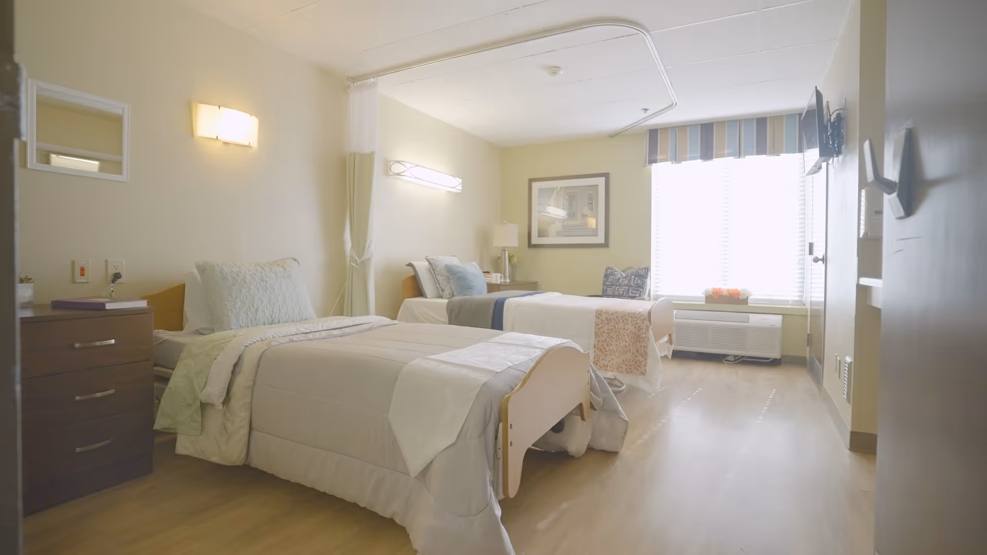 A bright and clean bedroom in a senior living facility with two single beds, each made with light-colored bedding and pillows. There is a nightstand with drawers between the beds, a wall-mounted light above each bed, a framed picture on the wall, and a large window with blinds and a striped valance letting in natural light. A flat-screen TV is mounted on the wall opposite the beds.