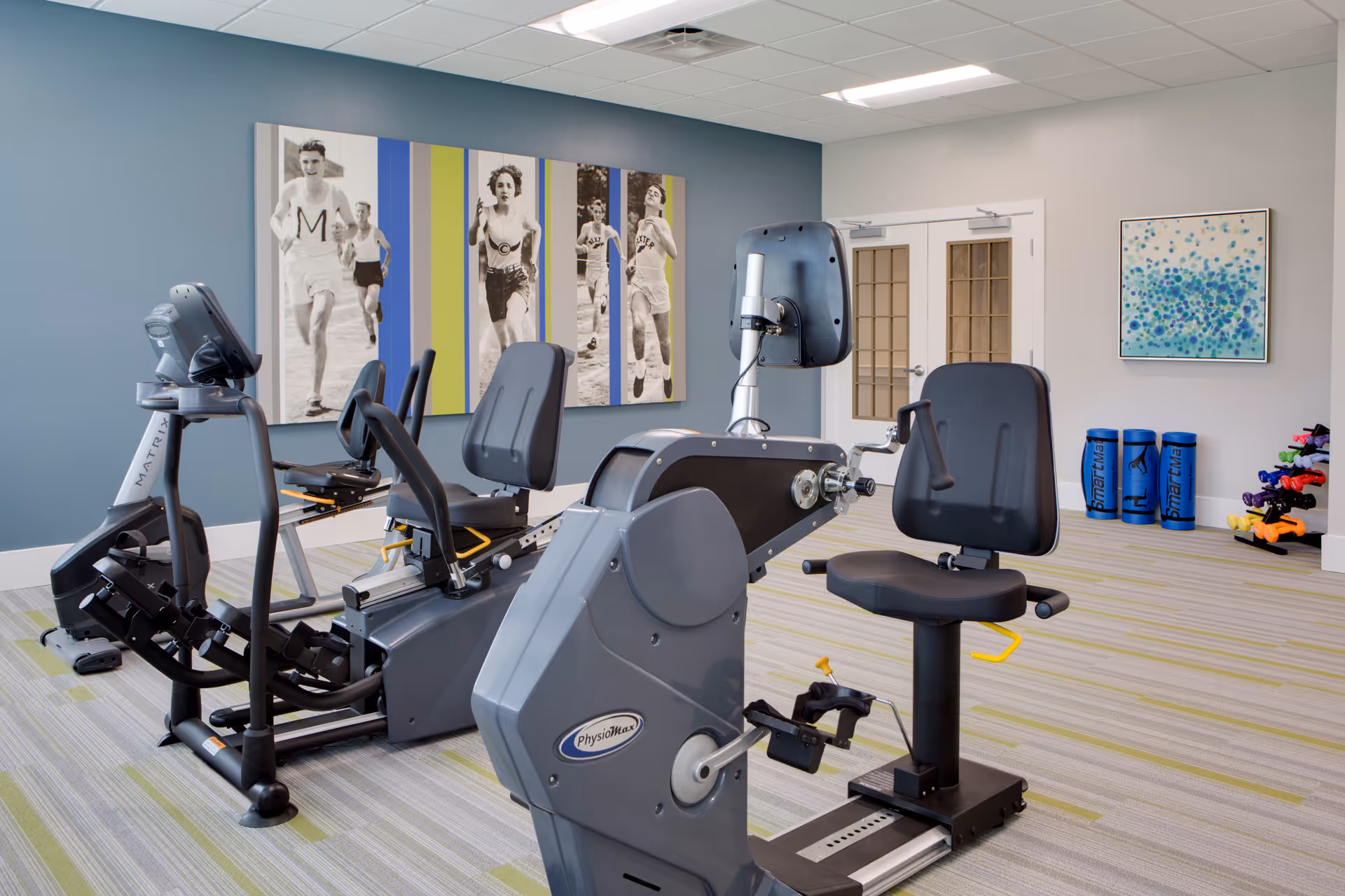 A fitness room with exercise equipment including recumbent bikes and a rowing machine. The room has a blue accent wall with a large vintage-style photo of runners, light-colored walls, and a patterned carpet. There are exercise mats and dumbbells neatly arranged in the corner.