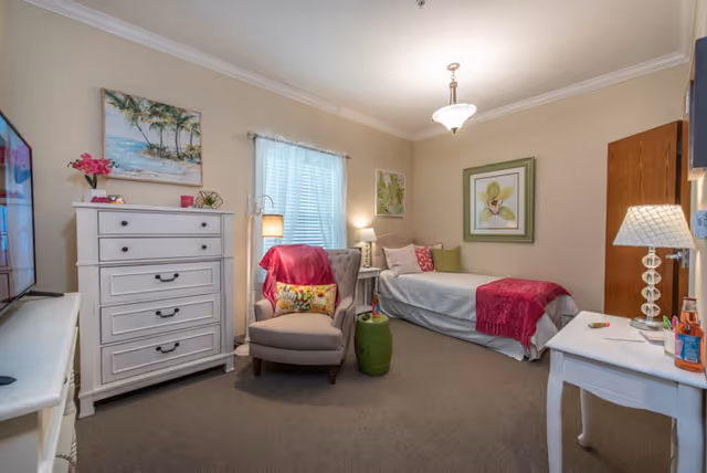 A cozy bedroom in an assisted living facility featuring a single bed with white and pink bedding, a gray armchair with a colorful pillow and pink throw, a white dresser with drawers, a white side table with a lamp, and framed artwork on beige walls. The room is softly lit with ceiling and table lamps, and a window with white blinds lets in natural light.