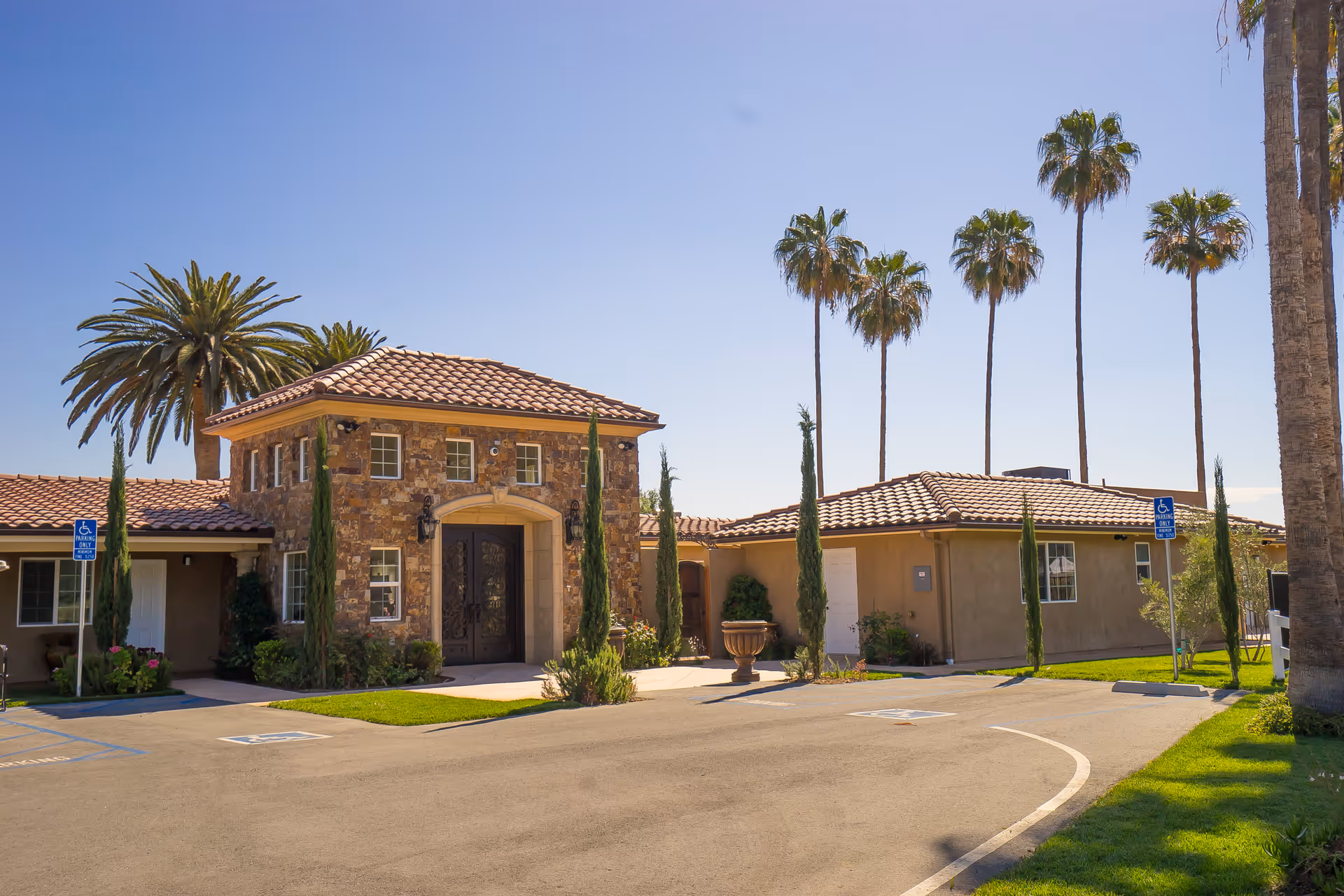 Exterior view of a single-story building with a stone facade entrance and tiled roof, surrounded by palm trees and small cypress trees. There are handicap parking spaces in front of the building under a clear blue sky.