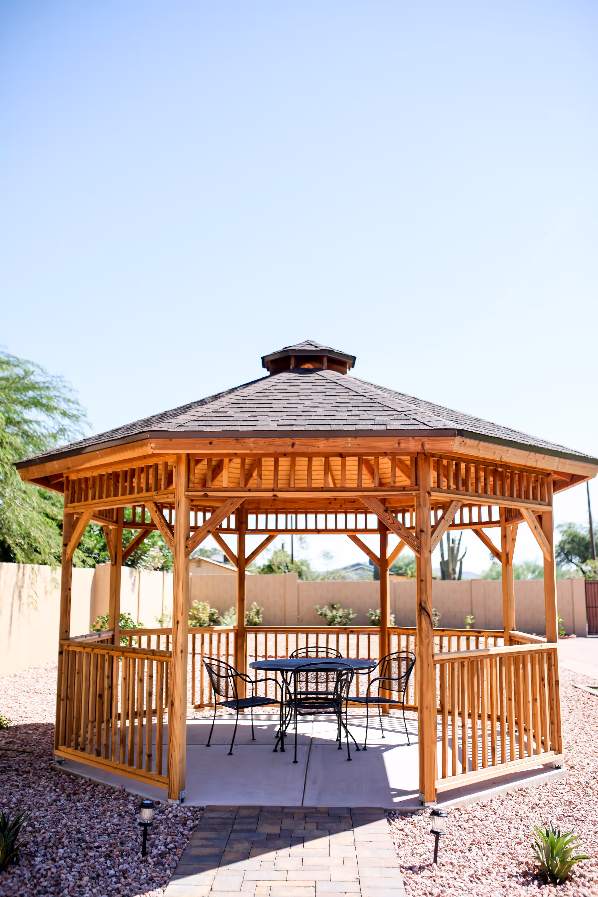 A wooden gazebo with a shingled roof situated outdoors on a paved area surrounded by gravel and desert landscaping. Inside the gazebo, there is a round metal table with four matching chairs. The sky is clear and blue.
