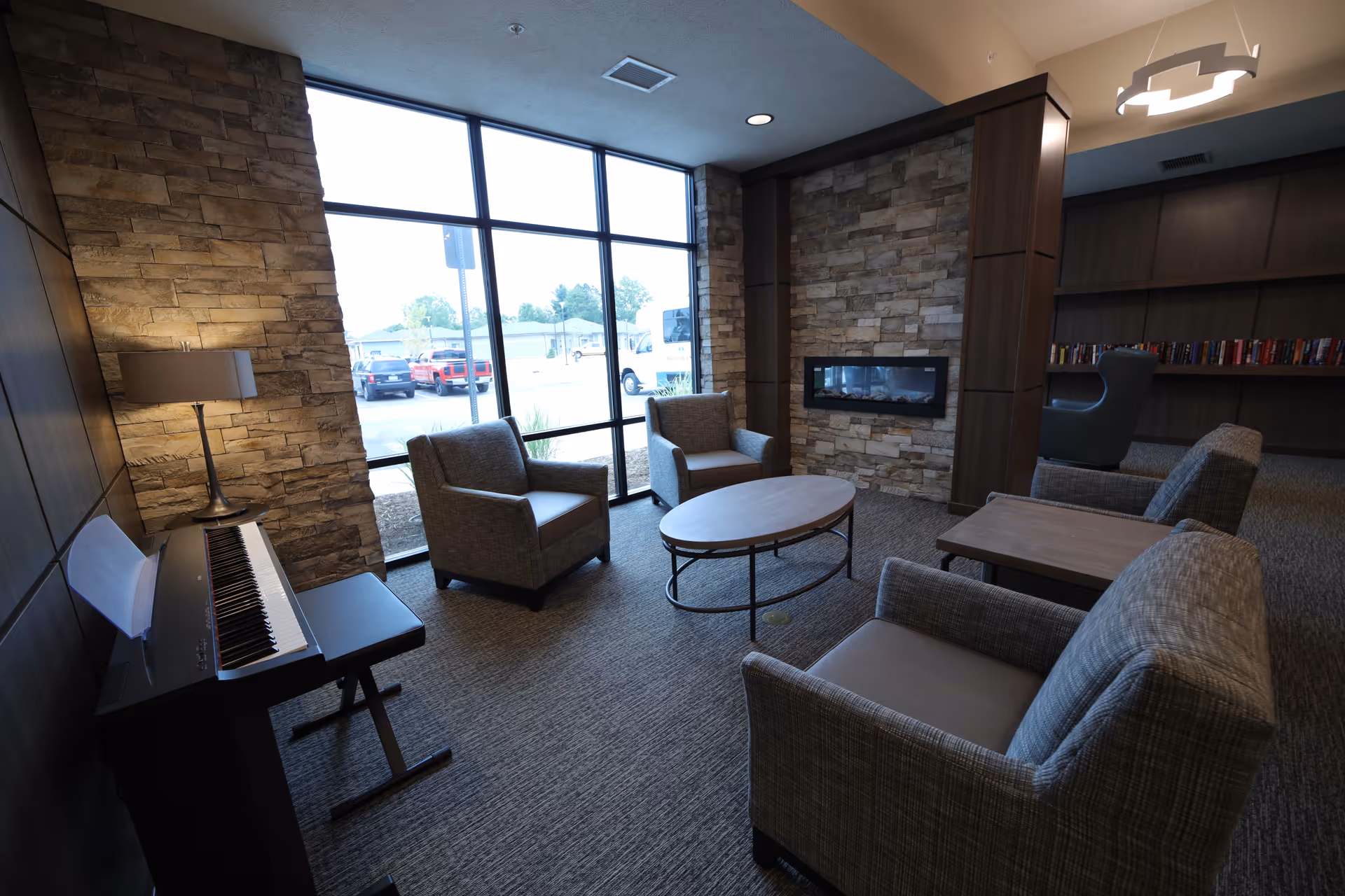 A comfortable lounge with armchairs around a round coffee table, a wall-mounted fireplace, a piano, and large windows looking out to the parking lot.